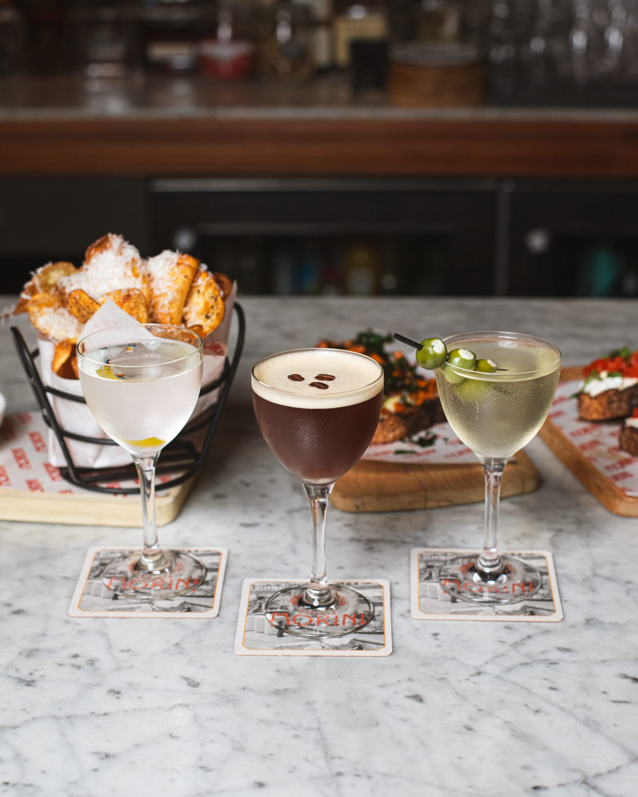 three martinis (classic, dirty, and espresso) in nick and nora glasses lined up in front of a basket of fresh, housemade rosemary chips, and bruschetta