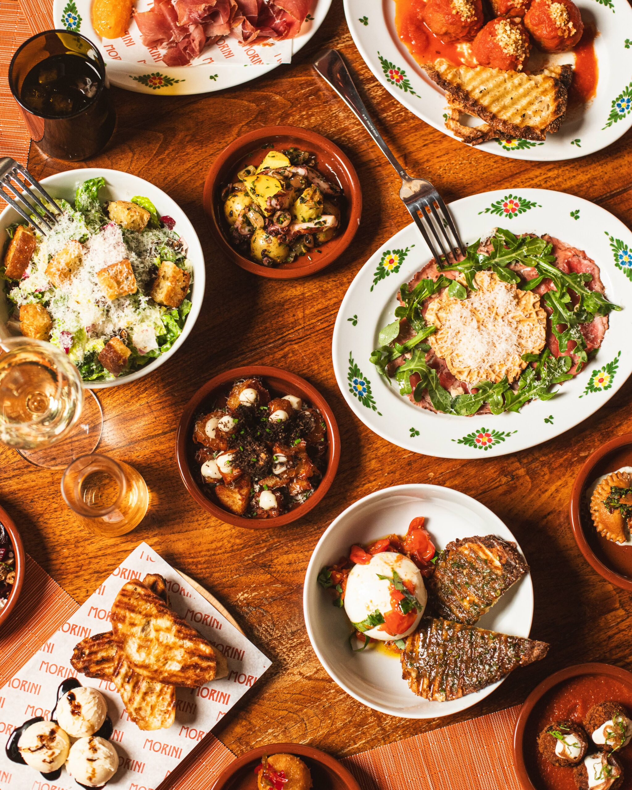 a group of northern italian dishes on a wooden table, including salads, burrata, meatballs, wine, and more.