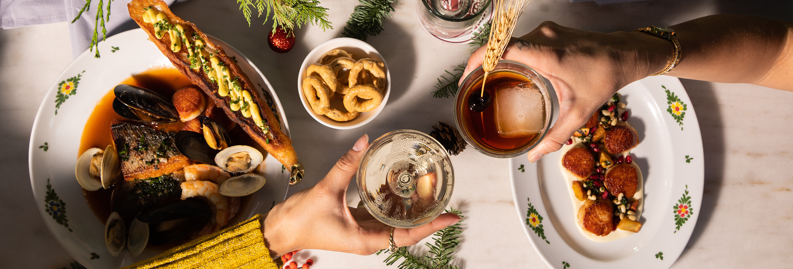 two hands cheers-ing cocktails over a table with seafood dishes and chrismas decorations