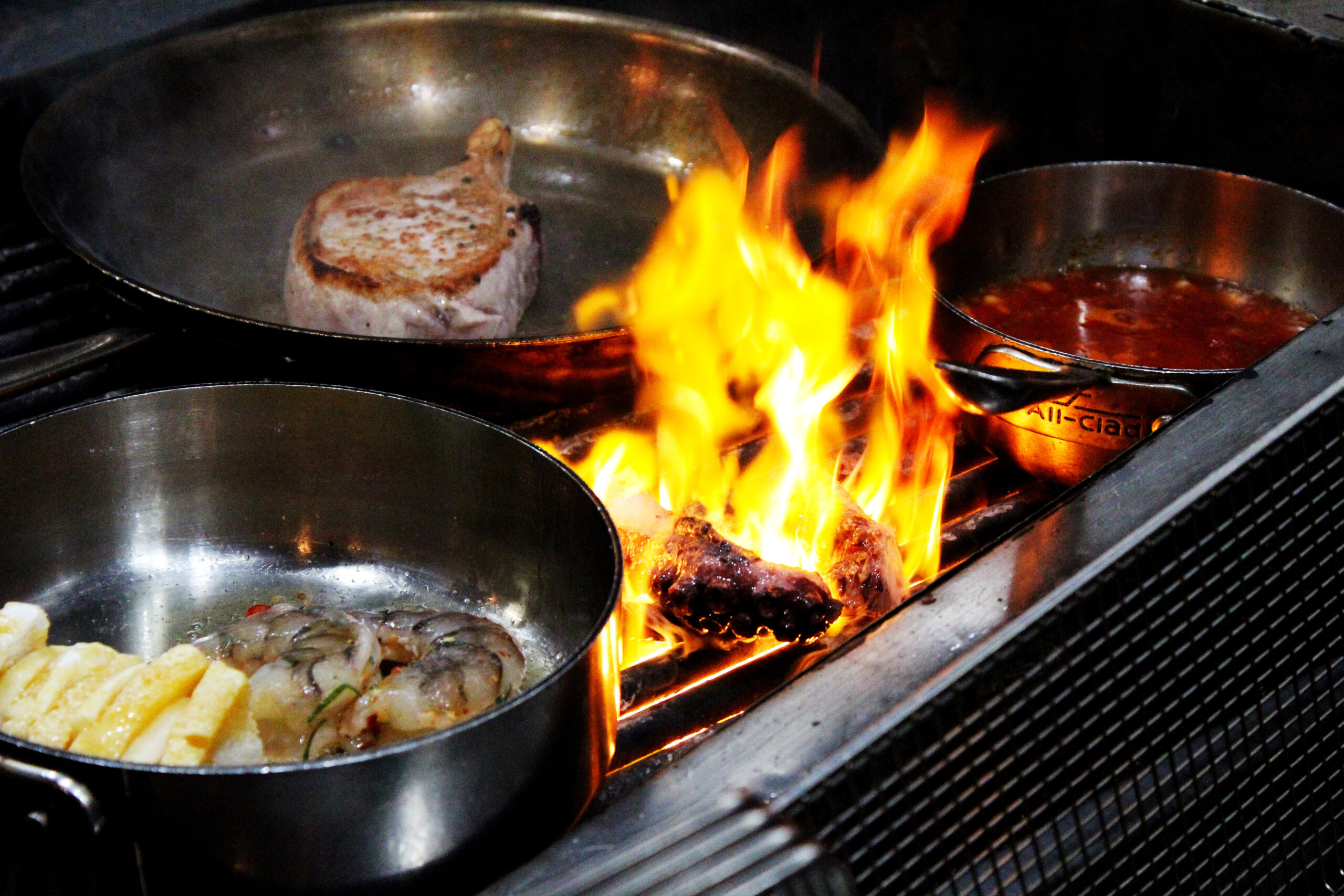 octopus flaming on the grill next to a pan searing pork, one with shrimp, and a pot with a red sauce