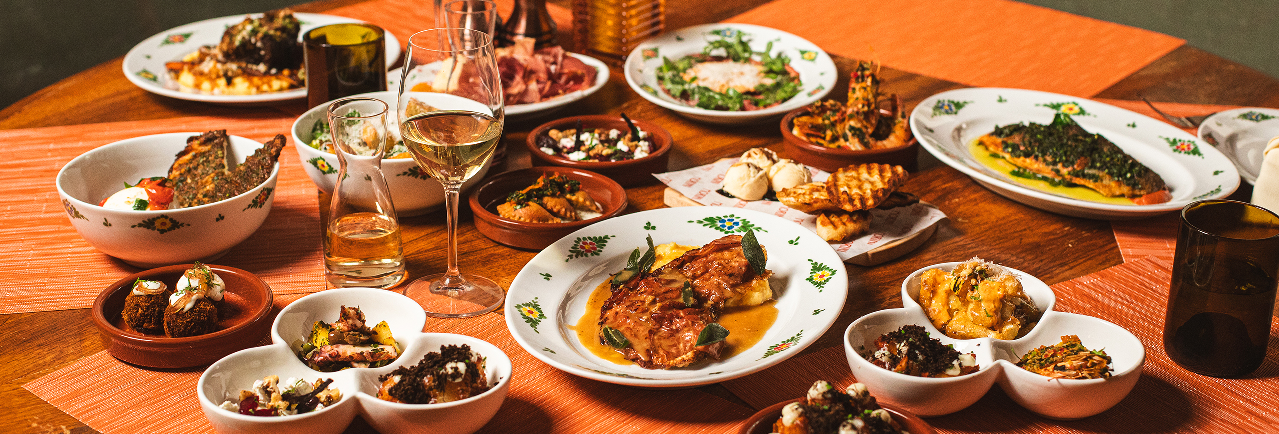 a group of northern italian dishes on a wooden table, including salads, burrata, meatballs, strattcotto, wine, and more.