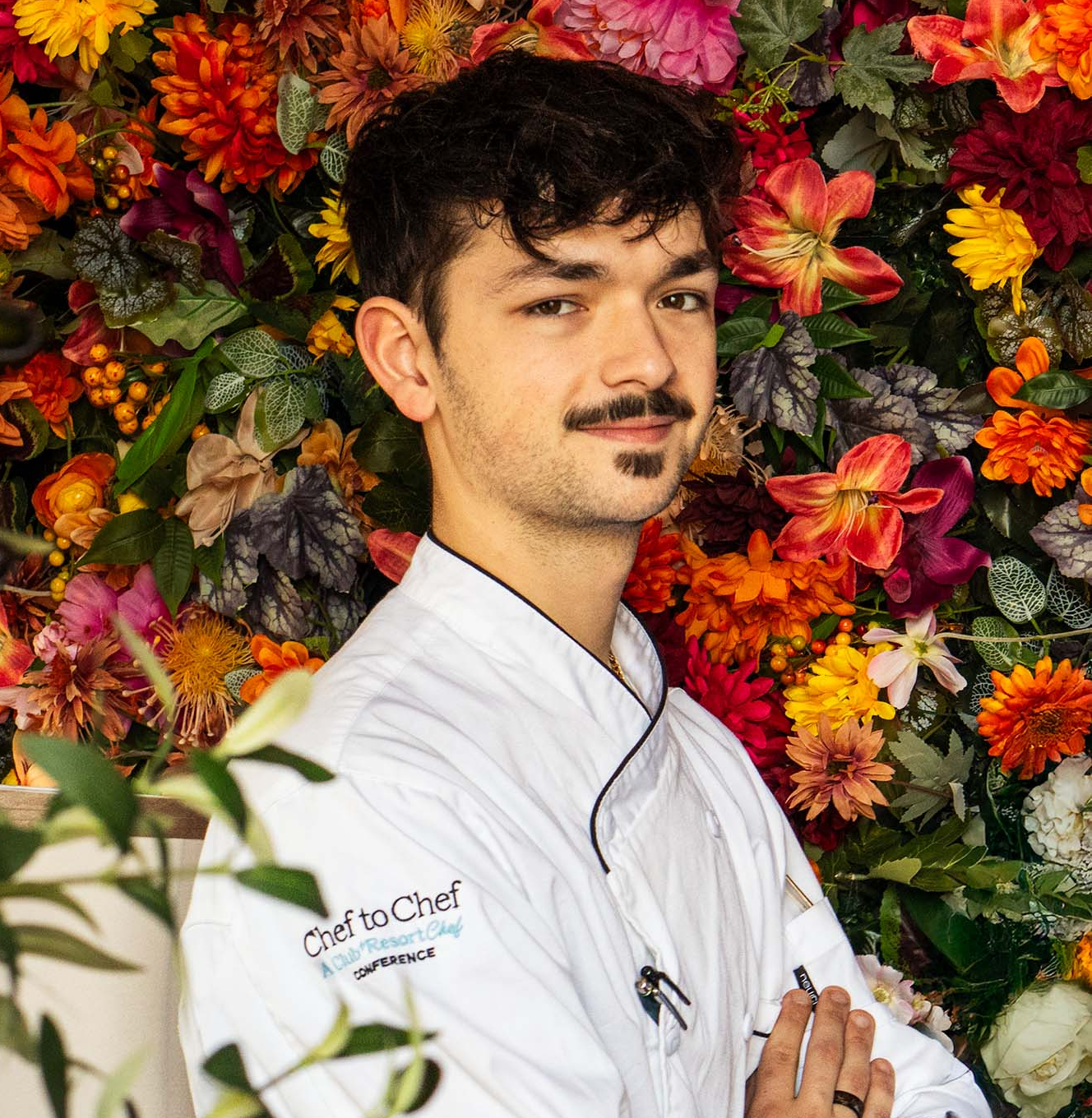 A young male chef with dark curly hair and a trimmed mustache stands confidently in front of a vibrant floral wall at Osteria Morini Bernardsville Restaurant. He wears a white chef’s coat embroidered with “Chef to Chef – A Club + Resort Chef Conference” on the sleeve and has his arms crossed, smiling subtly at the camera.
