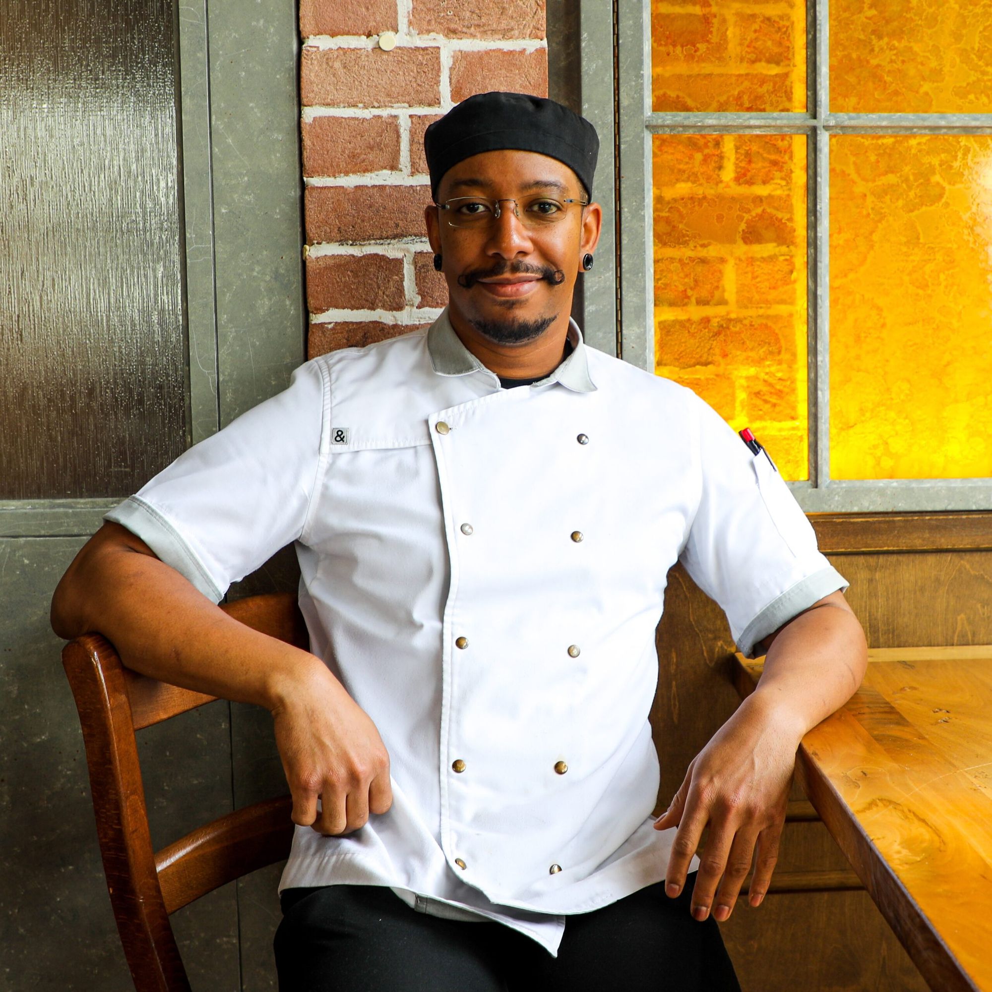 In chef's whites and a black cap, seated by a brick pillar and an amber glass window.