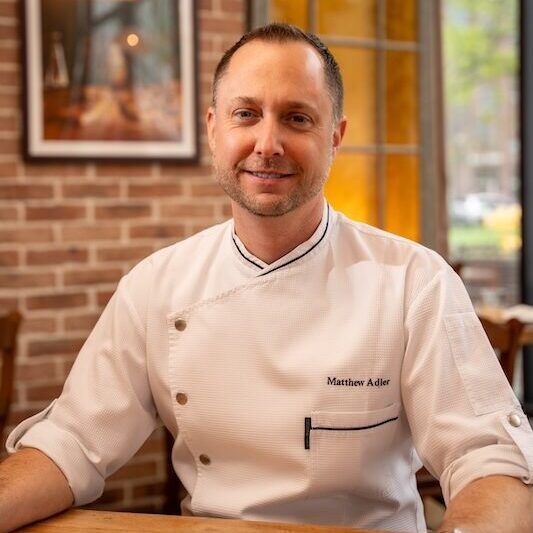 In chef's whites, seated at a table by an exposed brick wall.