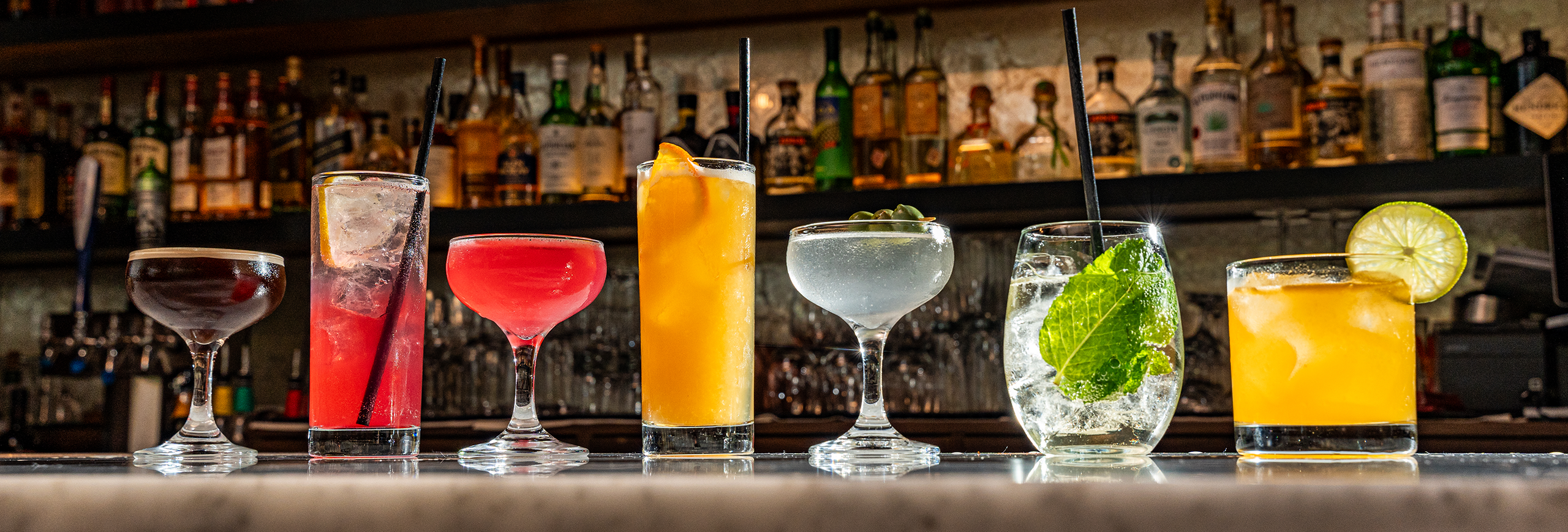 a line of cocktails on a bar top, all a variety of colors and in a variety of glasses