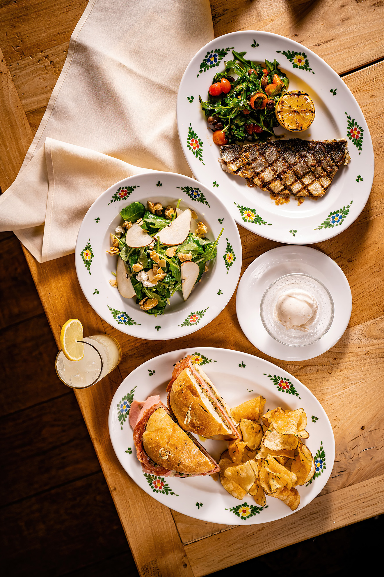 a top down view of a table with a salad, branzino, sandwich, drink, and a scoop of gelato.