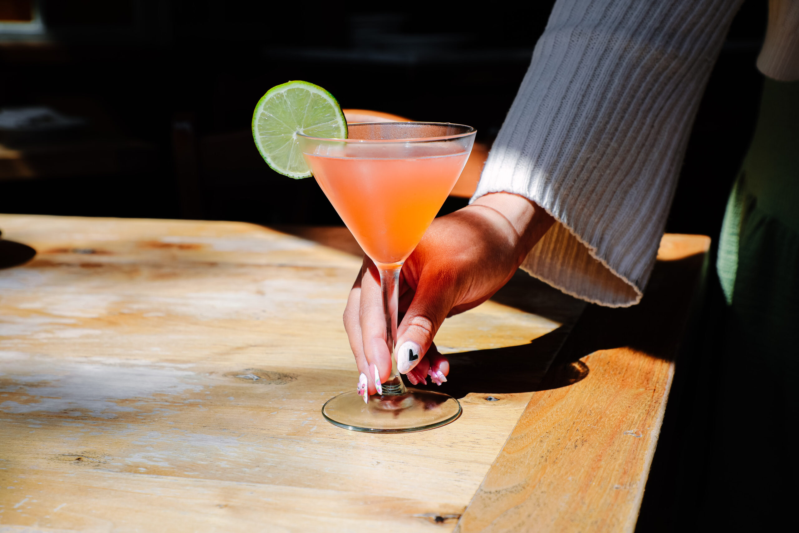 a hand holding a pink cocktail in a martini glass with a lime on the rim