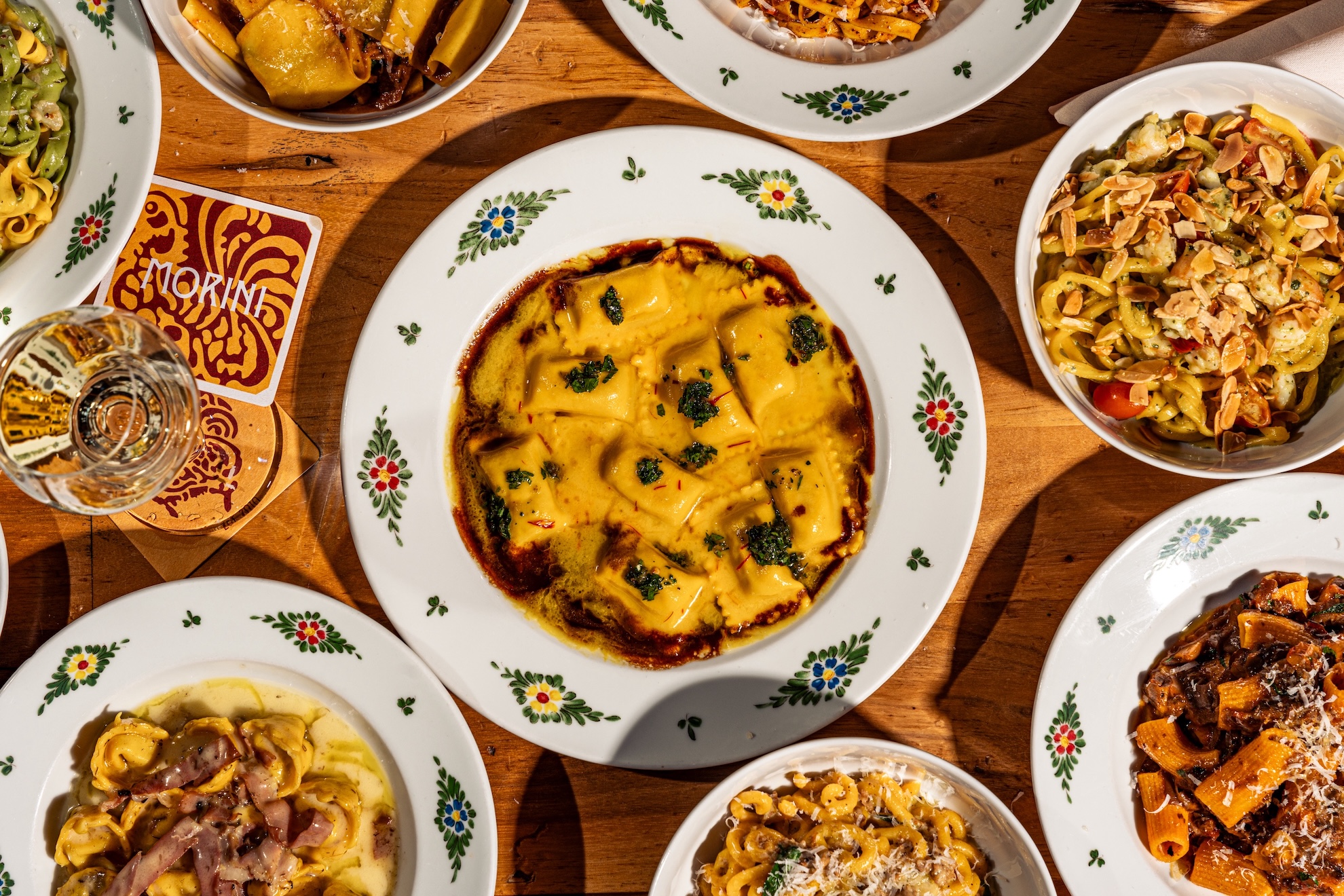 n overhead view of a wooden table filled with assorted Italian pasta dishes served in white plates decorated with small floral patterns; at the center is a saffron-colored ravioli dish with brown butter, surrounded by various pastas including rigatoni with ragu, tortellini in cream sauce, green tagliatelle, and a bowl topped with toasted almonds, plus a glass of white wine and Morini-branded coasters.